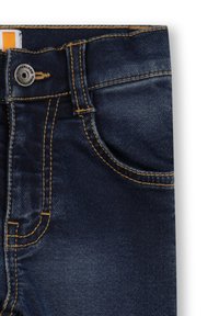 Primo piano di jeans in denim blu scuro che mostrano la tasca anteriore, il passante della cintura e il bottone di metallo con etichetta arancione e gialla all'interno della vita.