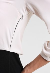 Rosa blus med långa ärmar och en figursydd passform. Har en sidoknäppning i svart som kontrasterar med det släta tyget.