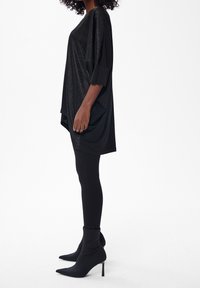 Femme portant une tunique noire ample et scintillante, des leggings noirs et des bottines noires à talons aiguilles pointus, debout de profil sur fond blanc.