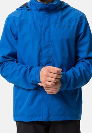 Homme portant une veste imperméable bleu vif avec des poignets élastiques, debout les mains jointes à la taille.