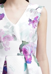 Robe fleurie avec un décolleté en V, présentant un tissu texturé blanc et des motifs floraux multicolores dans des tons de violet et de vert.