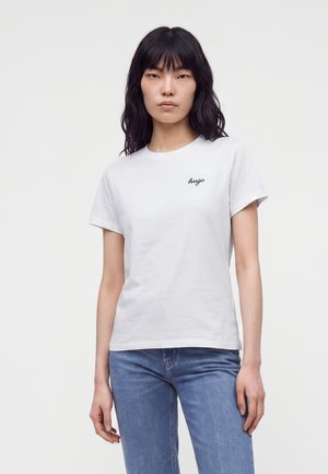Jonge vrouw met lang zwart haar, dragend een wit t-shirt met Hugo-logo en een blauwe spijkerbroek, staand tegen een effen lichte achtergrond.