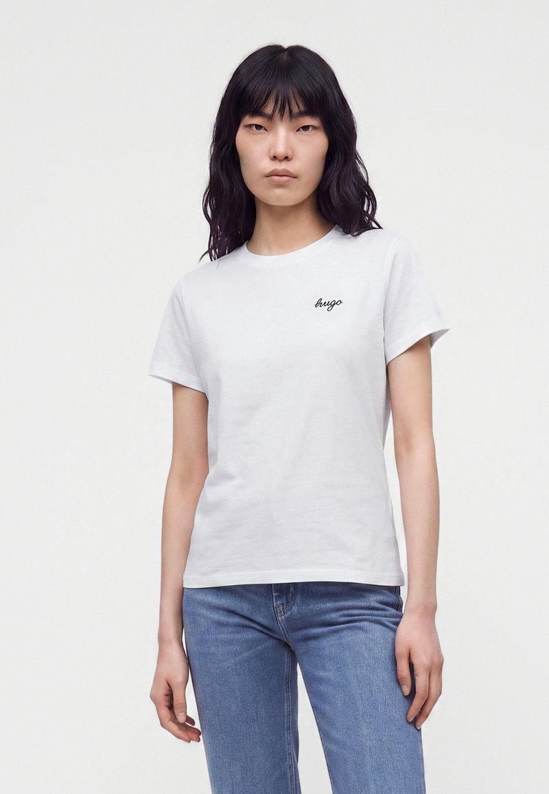 Jeune femme aux longs cheveux noirs, portant un t-shirt blanc avec le logo Hugo et un jean bleu, debout devant un fond clair et uni.