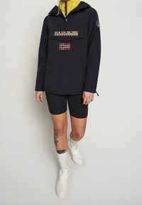 Giacca pullover blu scuro realizzata in materiale leggero, con tasca frontale, logo e dettaglio della bandiera. Abbinata a pantaloni corti neri e sneakers bianche.