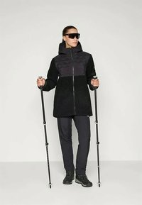 Jacheta neagră bicoloră cu material texturat în partea de jos, asortată cu pantaloni negri și bețe de drumeție. Ochelarii de soare completează ținuta.