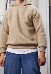 Beige Fleece-Kapuze mit strukturierter Oberfläche, verstellbarer Kordelzug-Kapuze und gerippten Bündchen, getragen über einem weißen T-Shirt und blauen Jeans.
