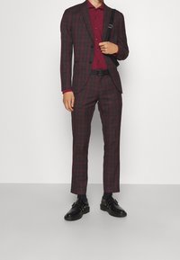 Traje de cuadros para hombre en burdeos con detalles en verde oscuro; cuenta con una chaqueta de dos botones, corte ajustado y una camisa a juego, combinado con zapatos negros.