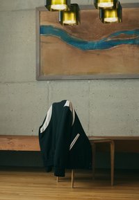 Black jacket with white accents, featuring a zippered front and elastic cuffs. Resting on a wooden bench against a textured concrete wall.