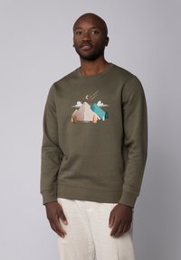 Un sweat-shirt vert olive avec un graphisme représentant une scène de montagne dans des couleurs pastel, col rond, manches longues, et poignets et ourlet côtelés.