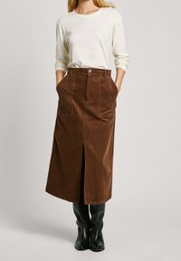 Gonna midi in corda marrone con spacco anteriore e tasche, abbinata a una maglietta a maniche lunghe color crema chiaro e stivali neri fino al ginocchio.