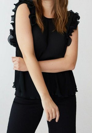 Kobieta w czarnej, bezrękawnikowej bluzce z falbanami i czarnych spodniach, z założonymi rękami, jedną ręką opartą na przeciwległym ramieniu.