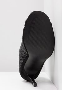 Zwarte hoge hak met een bovenwerk van gestructureerde stof, een gladde zwarte zool en een spits toelopende neus. Heeft een slank en gestroomlijnd silhouet.