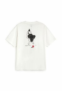 T-shirt blanc à manches courtes avec une illustration d’un chien noir et blanc regardant vers l’avant au-dessus d’une balle de tennis rouge dans le dos.