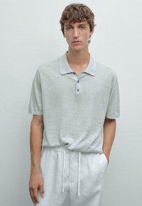 Světle šedé polo tričko s jemnou texturou, krátkými rukávy a límcem s třemi knoflíky, spárované se světle barevnými kalhotami na šňůrku.