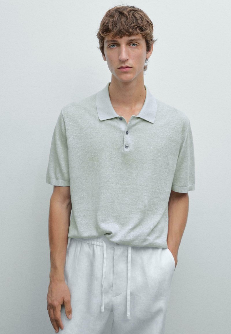 Světle šedé polo tričko s jemnou texturou, krátkými rukávy a límcem s třemi knoflíky, spárované se světle barevnými kalhotami na šňůrku.