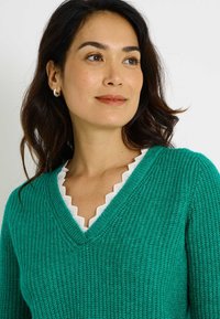 Suéter de cuello en V en un vibrante color verde azulado, con un patrón de tejido texturizado y un ribete blanco ondulado a lo largo del escote.