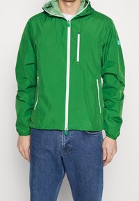 Giacca leggera verde con cappuccio, zip bianca e tasche con cerniera. Texture liscia, presenta una fodera a contrasto e un logo sulla manica.
