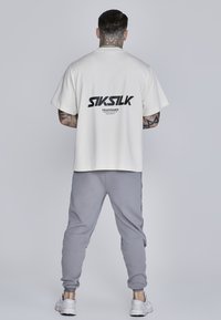 Balta kokvilnas T-kreklu ar melno "SIKSILK" logotipu aizmugurē, apvienots ar pelēkiem sporta biksēm un baltām kedām. Vienkārša fona.