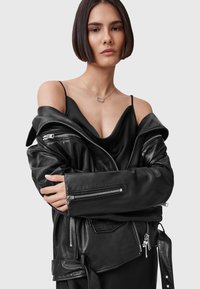 Zwarte leren jas met ritsen, over een zwart satijnen top. Model heeft kort, steil haar en draagt een zilveren ketting.