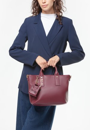 Bolso burdeos de piel texturizada, con herrajes dorados, asas dobles y una etiqueta de bolso mini extraíble sujeta con un llavero.