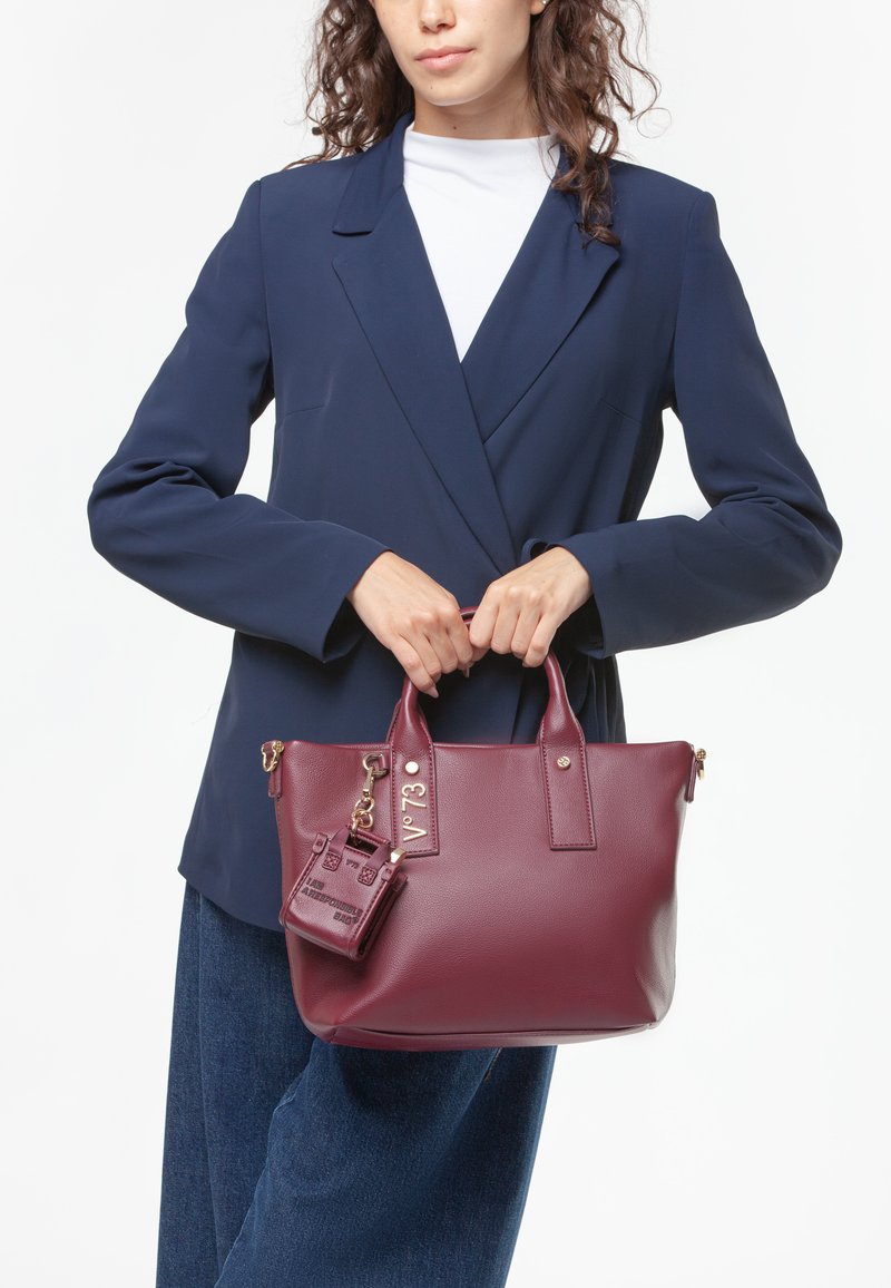 Bolso burdeos de piel texturizada, con herrajes dorados, asas dobles y una etiqueta de bolso mini extraíble sujeta con un llavero.