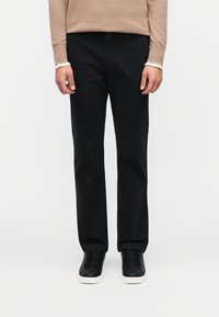 Μαύρο βαμβακερό παντελόνι με ίσια γραμμή, διαθέτει θηλιές για ζώνη, μπροστινές τσέπες και ομαλή υφή, συνδυασμένο με μαύρα αθλητικά παπούτσια.