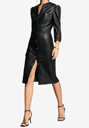 Woman wearing a black leather buttoned midi dress with puffed sleeves, chest pockets, and a front slit, paired with black high-heeled sandals.