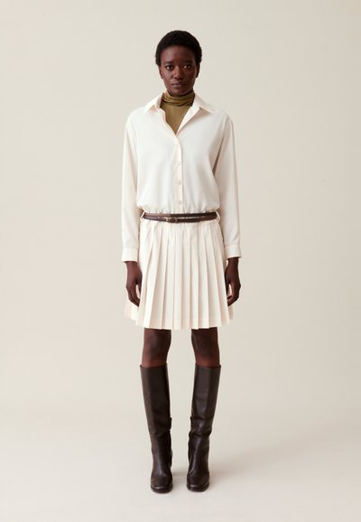 Femme portant une jupe plissée crème, une chemise blanche à boutons par-dessus un col roulé marron, une ceinture marron et des bottes brunes jusqu'aux genoux, se tenant contre un fond uni.