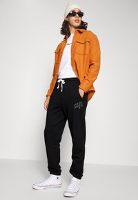 Camisa naranja de botones sobre una camiseta blanca, pantalones de chándal negros con logo y zapatillas altas blancas. Los accesorios incluyen un gorro beige y gafas de sol.