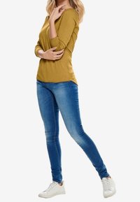 Senapsgul långärmad topp kombinerad med blå smala jeans och vita sneakers. Slät textur med åtsittande design, lätt drapering vid midjan.