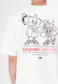 White cotton t-shirt featuring a black graphic of a smiling coffee pot and cup, with red text reading "Originals Coffee Club" and a logo.