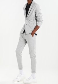 Traje gris claro con una tela suave y texturizada; incluye una chaqueta a medida y joggers ajustados. Combinado con zapatillas blancas y calcetines de caña corta.