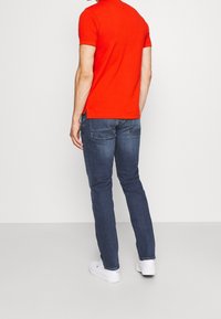 Camiseta roja de manga corta con bajo recto, combinada con jeans ajustados azul oscuro y zapatillas blancas. Detalles mínimos. Vista trasera.