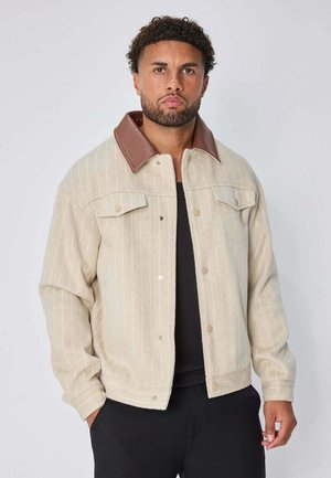 Homme portant une veste beige à rayures fines avec un col en cuir marron et une chemise noire, debout les mains dans les poches devant un fond uni.