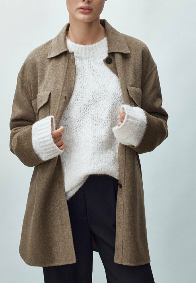 Manteau en mélange de laine marron avec de grandes poches avant, fermeture à boutons, et un pull blanc doux en-dessous. Le modèle porte un pantalon foncé. Fond clair.