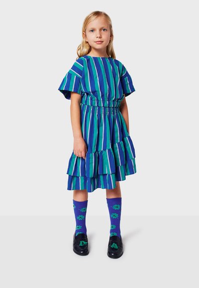 Blaue und grüne gestreifte Kleid mit kurzen Ärmeln, Gummizug in der Taille und gerüschtem Saum. Kombiniert mit lila Socken mit Blumenmuster und schwarzen Schuhen.