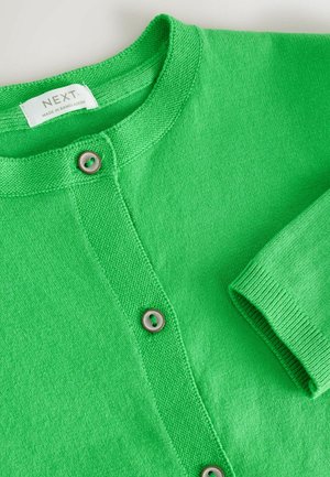 Grøn strikket cardigan med rund halsudskæring, synlige knapper og en påsyet label med teksten "NEXT Made in Bangladesh."