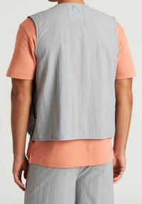 Gilet senza maniche grigio chiaro in tessuto strutturato, indossato sopra una camicia a maniche corte color corallo, con una vestibilità comoda e dettagli cuciti.