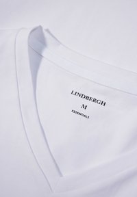 Biała bawełniana koszulka z dekoltem w serek. Posiada metkę z napisem "LINDBERGH M ESSENTIALS" w czarnym kolorze. Gładka tekstura.