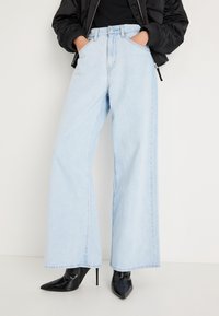 Φαρδιά τζιν από denim σε ανοιχτό μπλε χρώμα, με ψηλή μέση, μπροστινές τσέπες και καθαρό φινίρισμα στο τελείωμα, συνδυασμένα με μαύρα ψηλοτάκουνα παπούτσια.