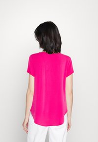 Haut à manches courtes et col rond en tissu rose vif. Texture lisse avec un ourlet arrondi. Porté avec un pantalon blanc, affichant une coupe décontractée.