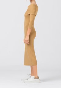 Abito midi in maglia, a spalle scoperte, color nocciola, con texture a coste. Maniche lunghe, silhouette aderente, abbinato a sneakers bianche con dettagli color nocciola.