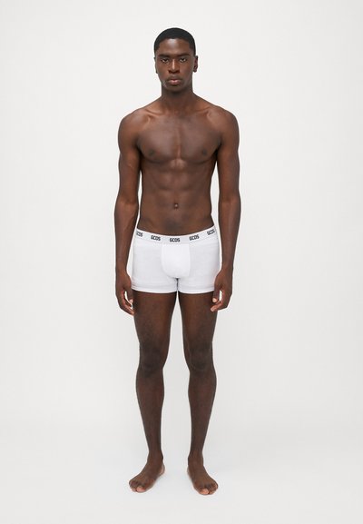 Witte katoenen boxershorts met een zwarte logo tailleband. Model staat met blote voeten, toont een strakke pasvorm, gladde textuur en minimale bedekking.