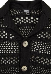 Juodas megztas kardiganas su audiniu raštu, turintis apykaklę ir dvi mygtukus. Pagamintas iš tekstūruotos medžiagos, kuri rodo skaidrų dizainą.