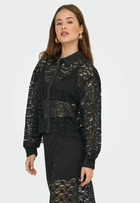 Blouse en dentelle noire avec un col, fermeture à boutons et manches longues. Présente des motifs floraux et un design court et superposé.