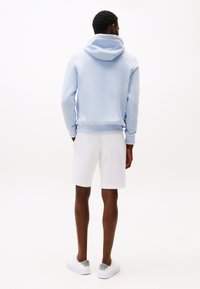 Hanorac albastru deschis cu glugă mare, asortat cu pantaloni scurți albi și adidași albi. Material neted și manșete tivite. Design minimalist.