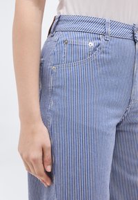 Πλευρικό μέρος κορμού και χέρι ατόμου που φορά μπλε και λευκά τζιν με κάθετες ρίγες και λευκό τοπ πάνω σε απλό φωτεινό φόντο.