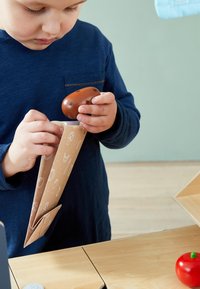 Bruine papieren tas met speelse illustraties, vastgehouden door een kind, met daarin een houten voedselitem. Gladde textuur en zachte kleuren.