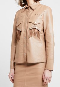 Läderjacka i brun färg med fransdetaljer på bröstfickorna, silvernitar längs axlarna, knäppning fram och långa ärmar. Slät textur.