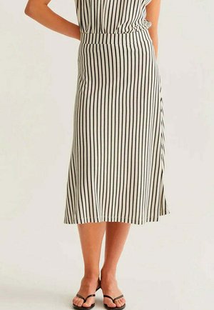 Mujer con falda midi de rayas verticales blancas y negras con una abertura, combinada con sandalias negras con tiras, de pie con las manos detrás de la espalda.